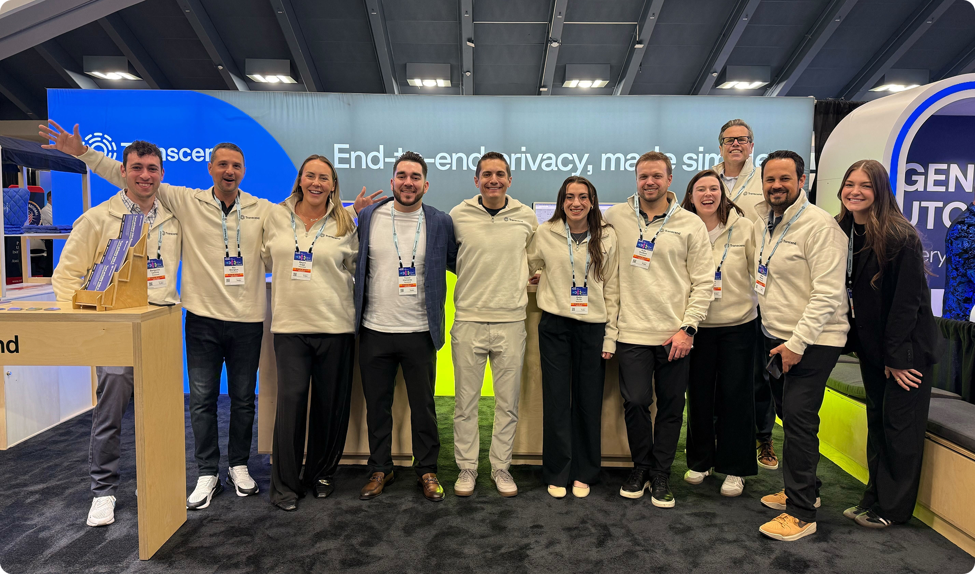 A group of people wearing matching light-colored sweaters stands together, smiling, in front of a booth with a blue and gray backdrop displaying the text “End-to-end privacy, made simple.”