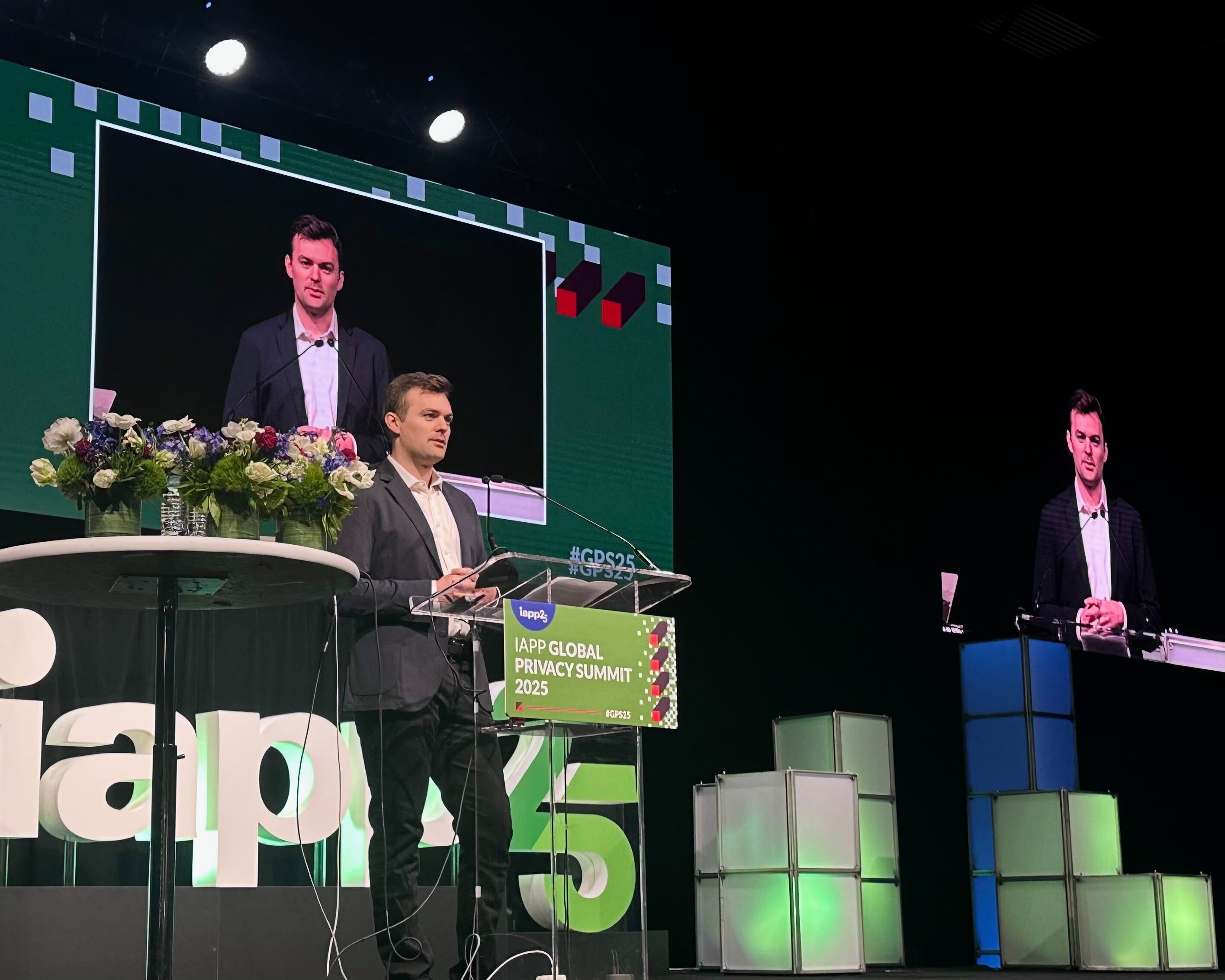 A speaker stands at a podium on stage during the IAPP Global Privacy Summit 2025, with a large screen displaying his image in the background.
