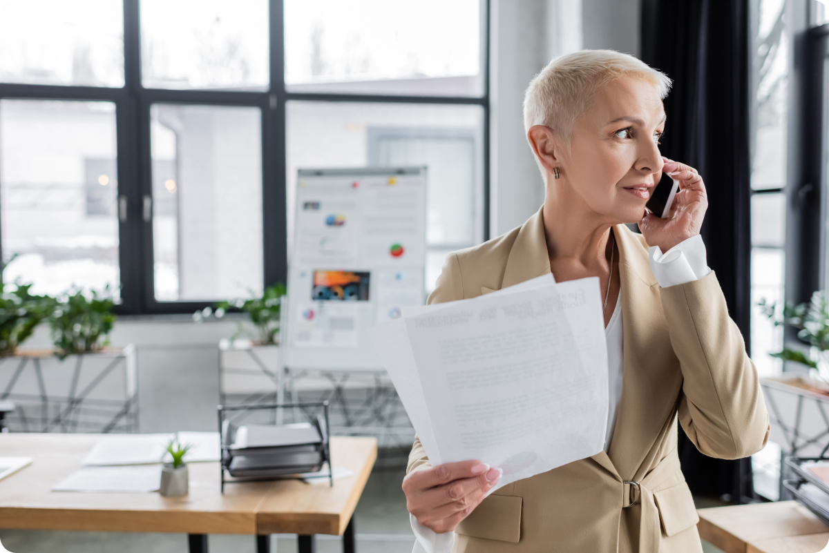 Experienced professional woman on the phone looking out the window of office holding documents