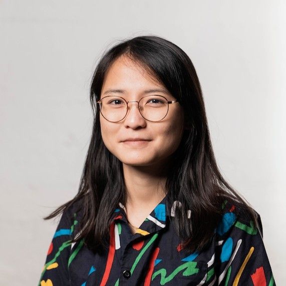 A person with long dark hair and glasses is wearing a colorful, patterned shirt and smiling softly against a light background.