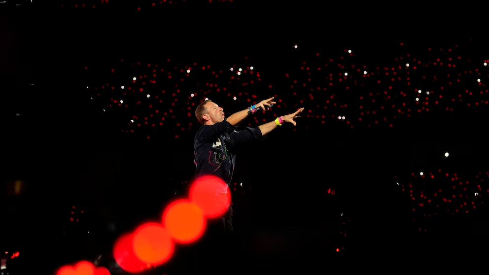 A performer on stage gestures energetically towards the audience, surrounded by a sea of red and white lights in the dark background.