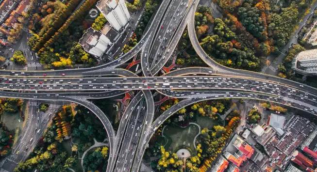 Aerial view of a complex multi-level highway interchange surrounded by dense urban buildings and greenery, with numerous vehicles traveling on the roads.