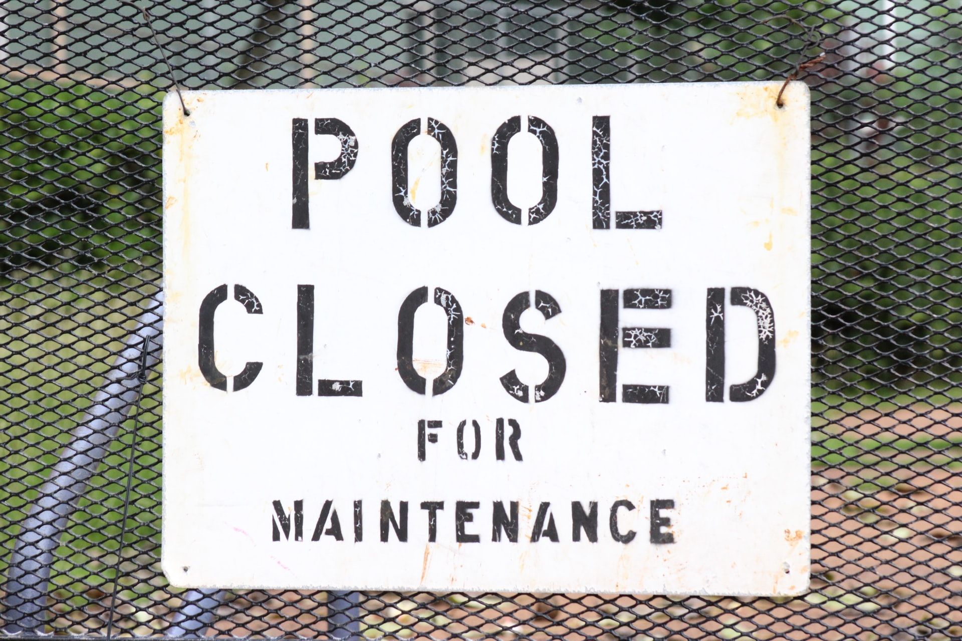 A sign on a metal mesh fence reads "POOL CLOSED FOR MAINTENANCE" in large black letters.