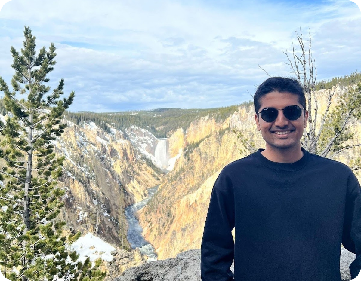 A person wearing sunglasses and a black sweater smiles in front of a scenic view featuring a waterfall cascading into a canyon, surrounded by pine trees and a cloudy sky.