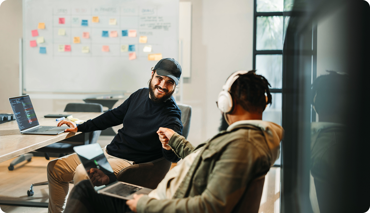 Two people sit at a table with laptops, smiling and fist-bumping, with a whiteboard covered in colorful sticky notes in the background. One person wears headphones and the other a cap.