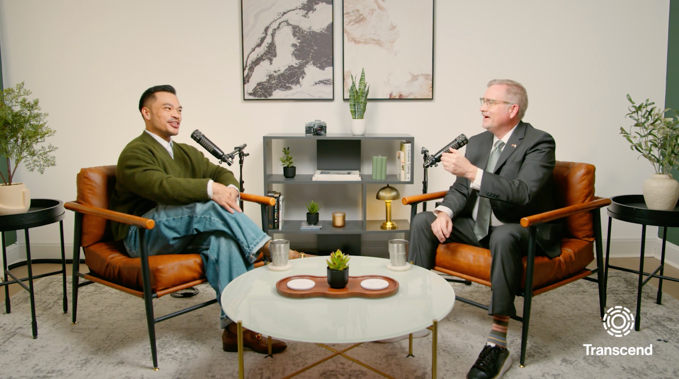 Two men are sitting in orange chairs, engaged in conversation, each holding a microphone. The setting is a warmly lit room with a modern decor featuring a table, a bookshelf with plants, and artwork on the walls.
