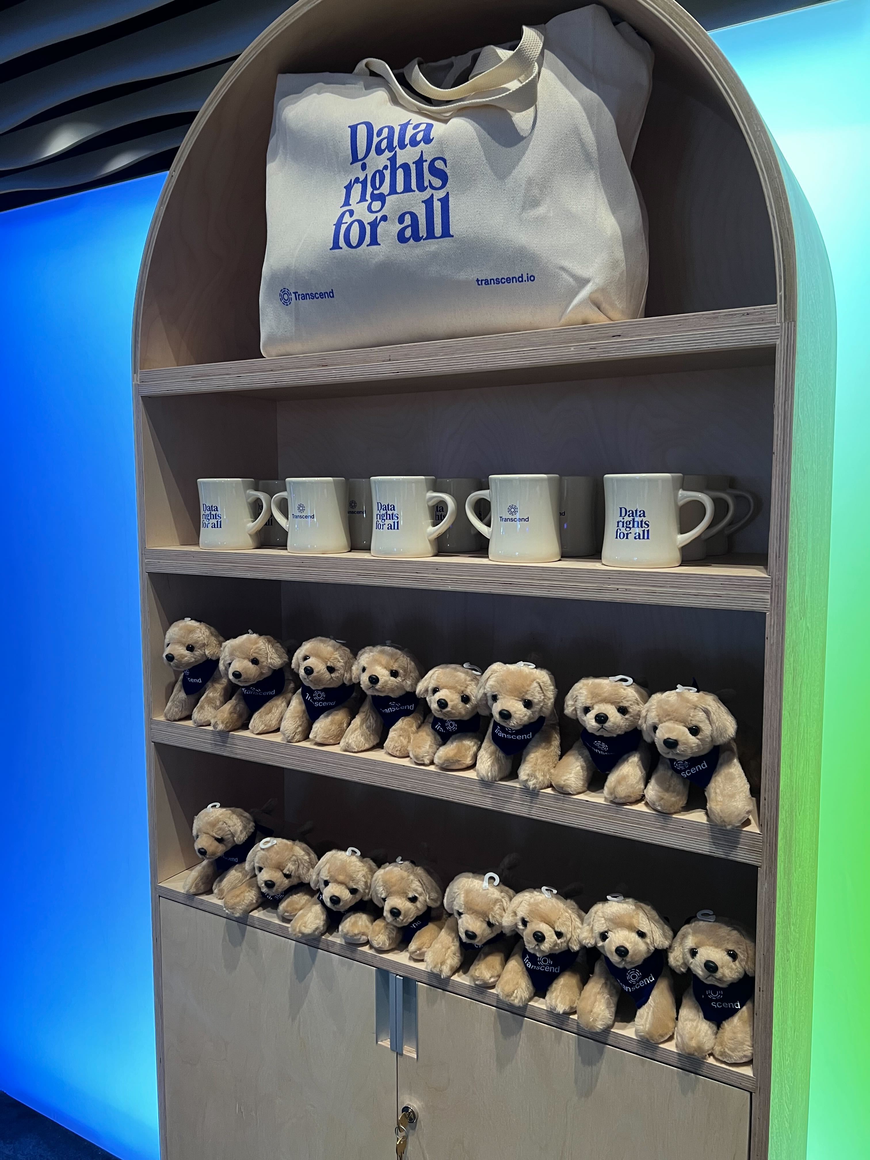 A wooden shelf displays a canvas bag with "Data rights for all" printed on it, mugs, and two rows of small teddy bears. The background features a gradient of blue and green lighting.