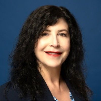 A woman with long, wavy dark hair smiles while wearing a dark blazer against a blue background.