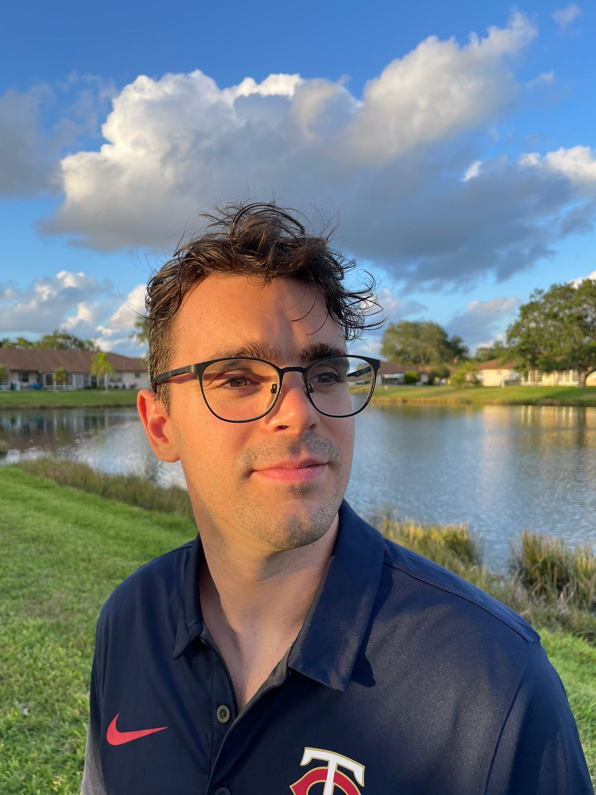A person with glasses stands by a lake under a blue sky with clouds, wearing a navy shirt featuring a logo.