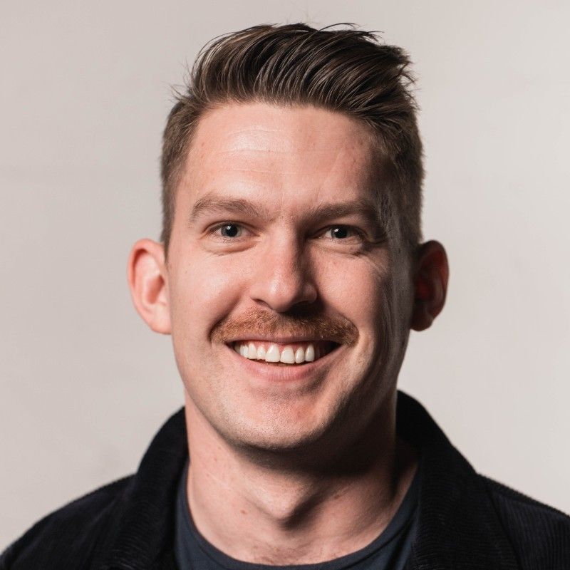 A man with short hair and a mustache is smiling, wearing a dark jacket and a t-shirt, set against a plain background.