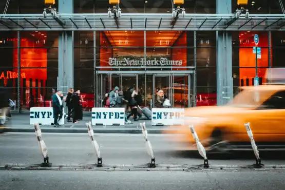 People are gathered outside the entrance to The New York Times building, with a yellow taxi blurred in motion passing by in front. NYPD barriers are positioned along the street.