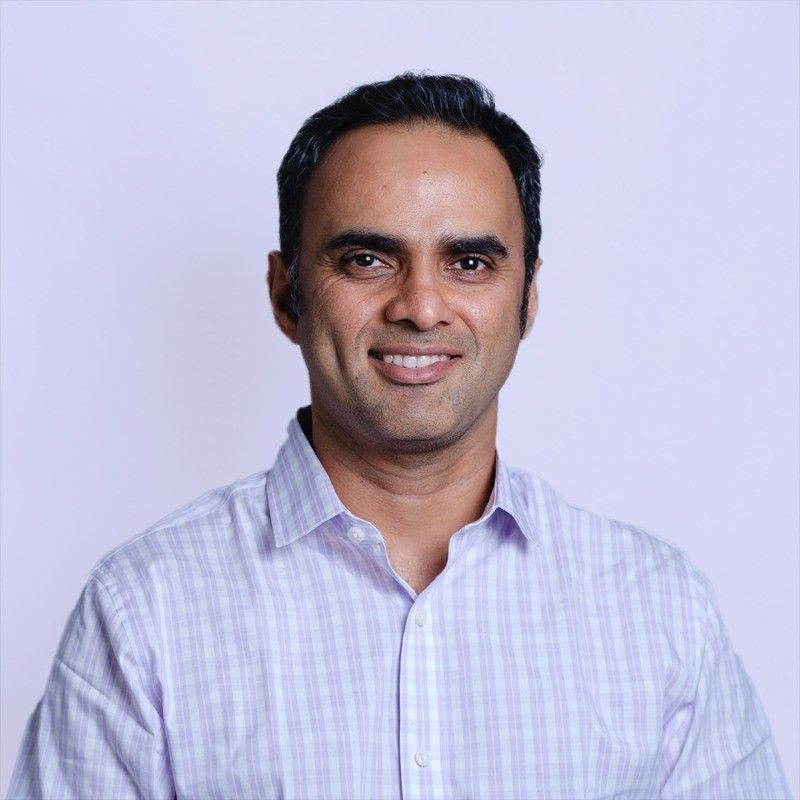 A smiling man with short dark hair wearing a light purple checkered shirt against a plain light background.