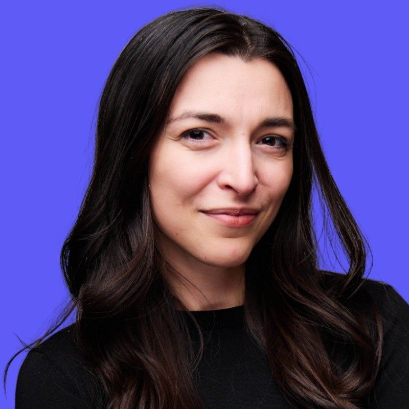 A person with long, dark hair and a slight smile against a solid purple background.