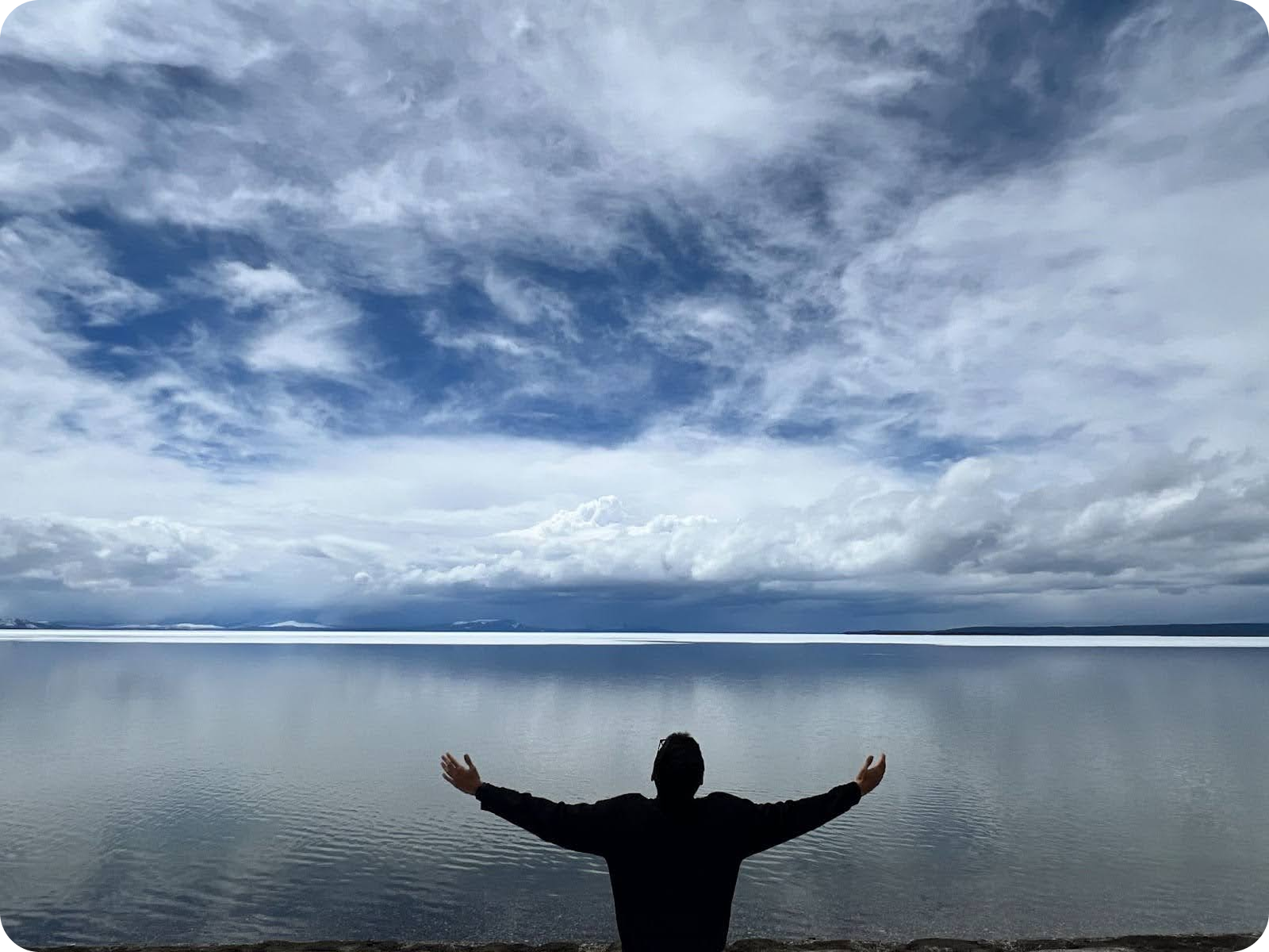 A person stands with arms outstretched, facing a vast, calm body of water under a dramatic, cloudy sky.