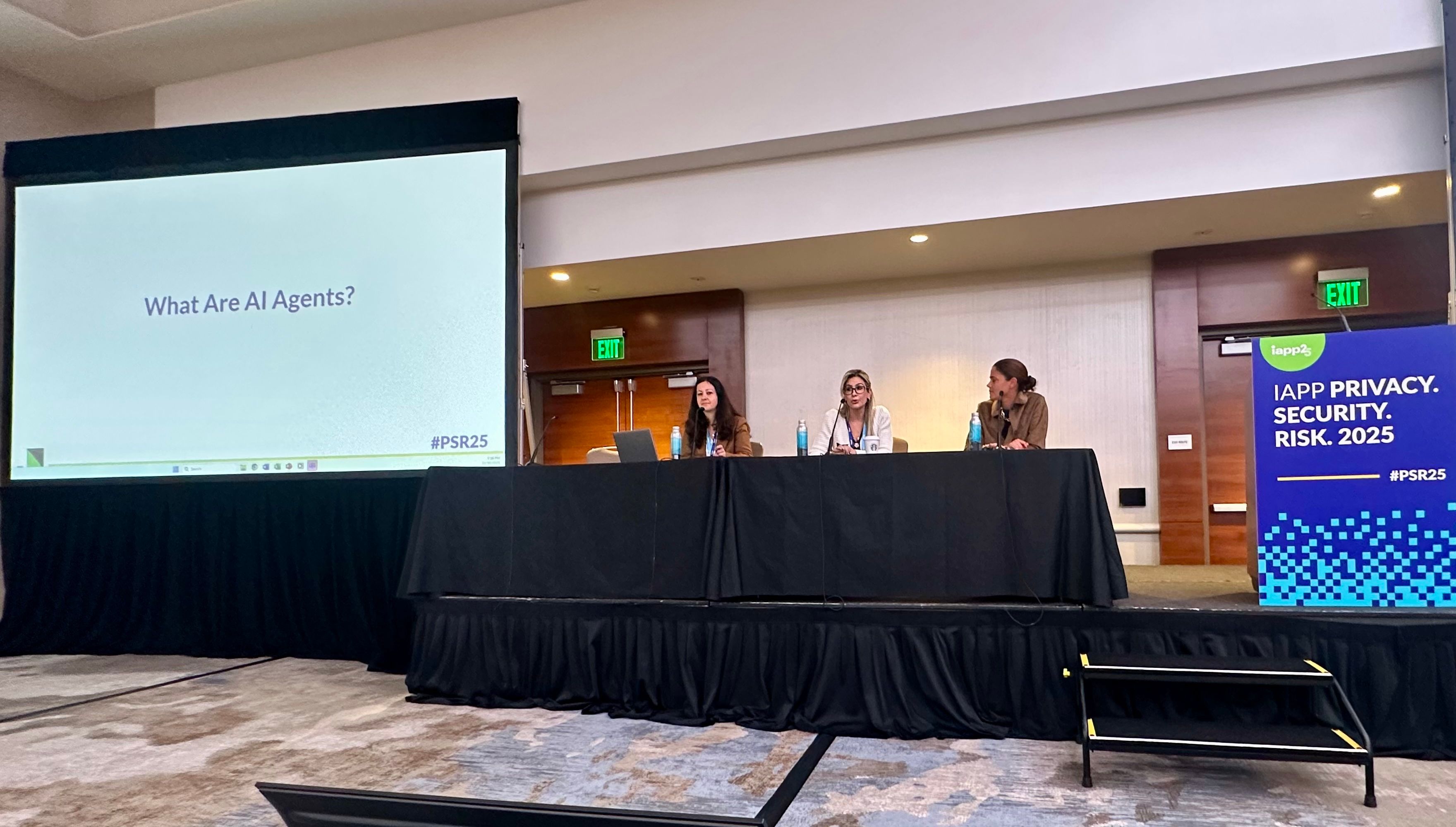 Three people sit at a panel discussion table with a large screen next to them displaying the text "What Are AI Agents?" and a conference banner reading "IAPP Privacy. Security. Risk. 2025" in the foreground.