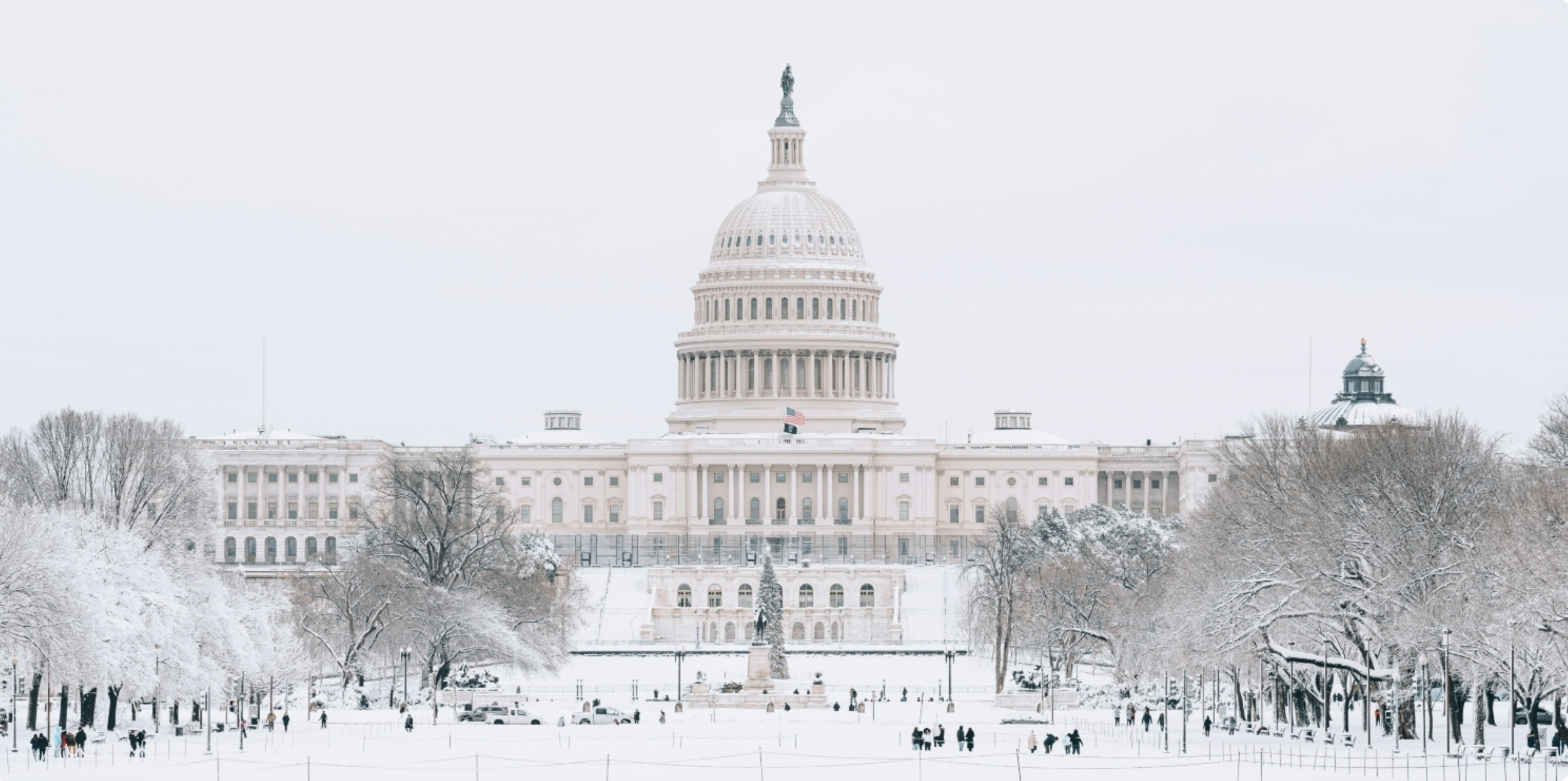 The U.S. Capitol building covered in snow, surrounded by snow-laden trees and a few people walking around.