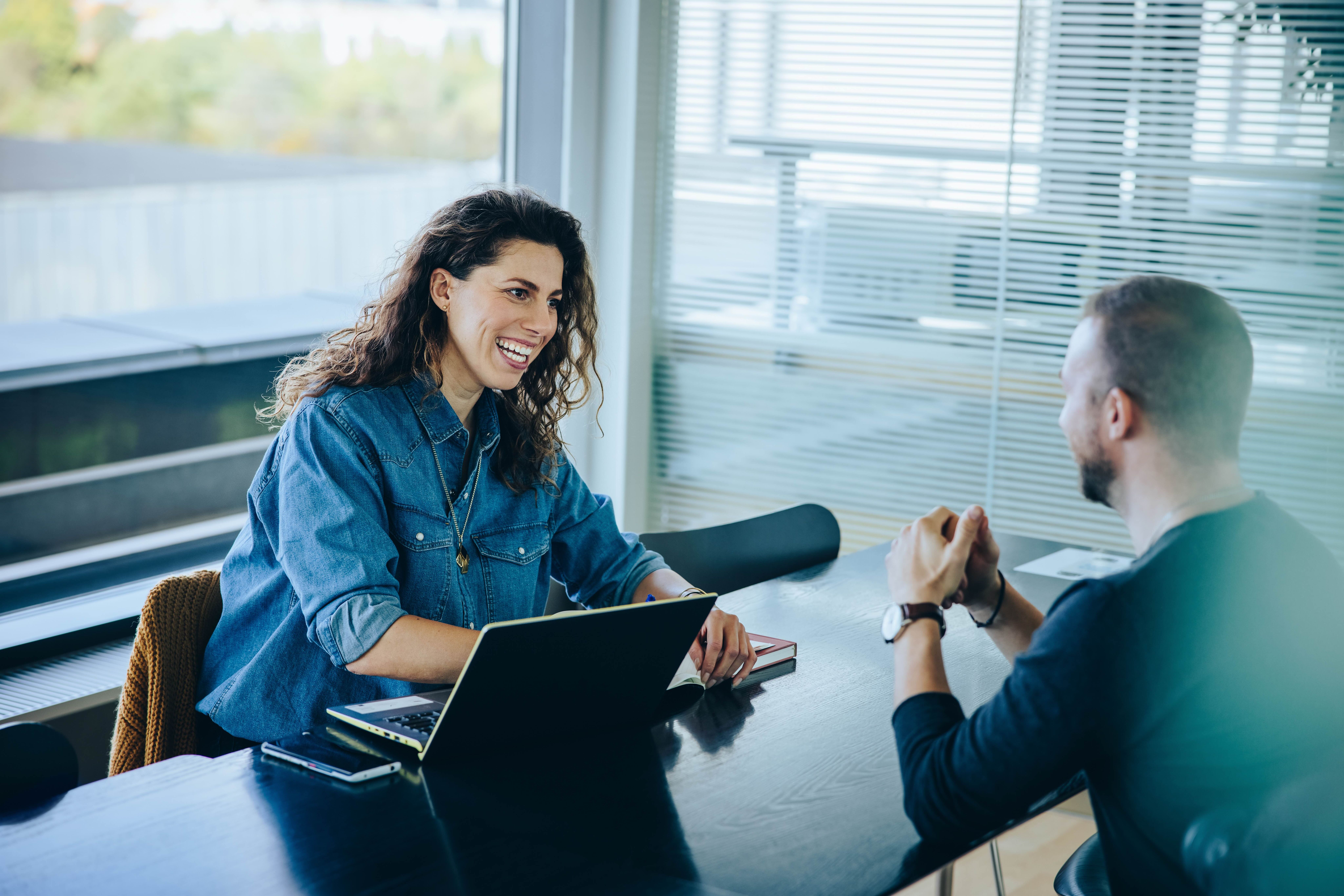 A smiling woman with a laptop talks to a man across an office table.