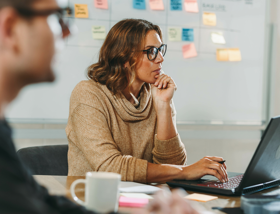 A woman with glasses and a tan sweater sits at a table, focused on her laptop. A whiteboard with colorful sticky notes is blurred in the background.