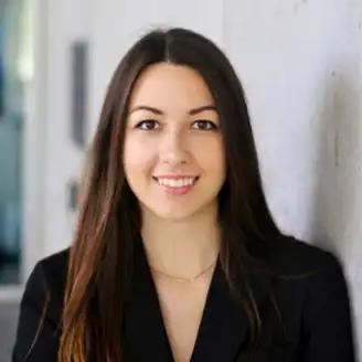 A person with long dark hair and a black blazer smiles while standing against a light-colored wall.