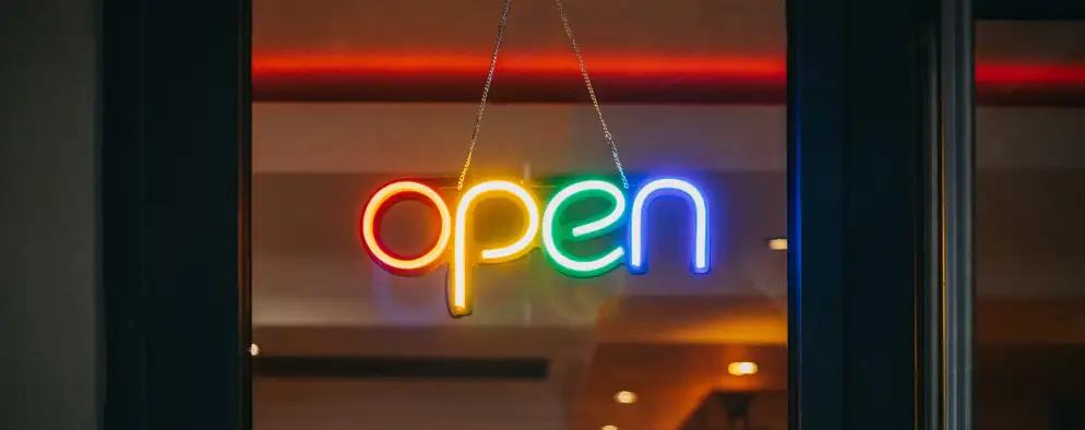 A brightly lit, multicolored neon sign displaying the word "open" hangs in a window against a warmly lit indoor backdrop.