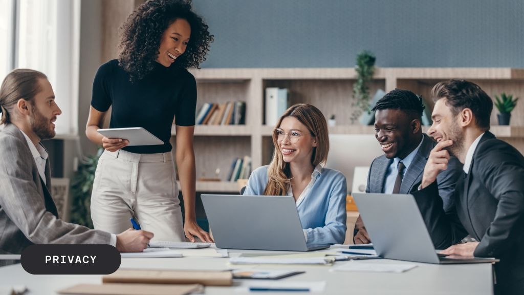 A diverse group of five smiling professionals meeting in a modern office with laptops.
