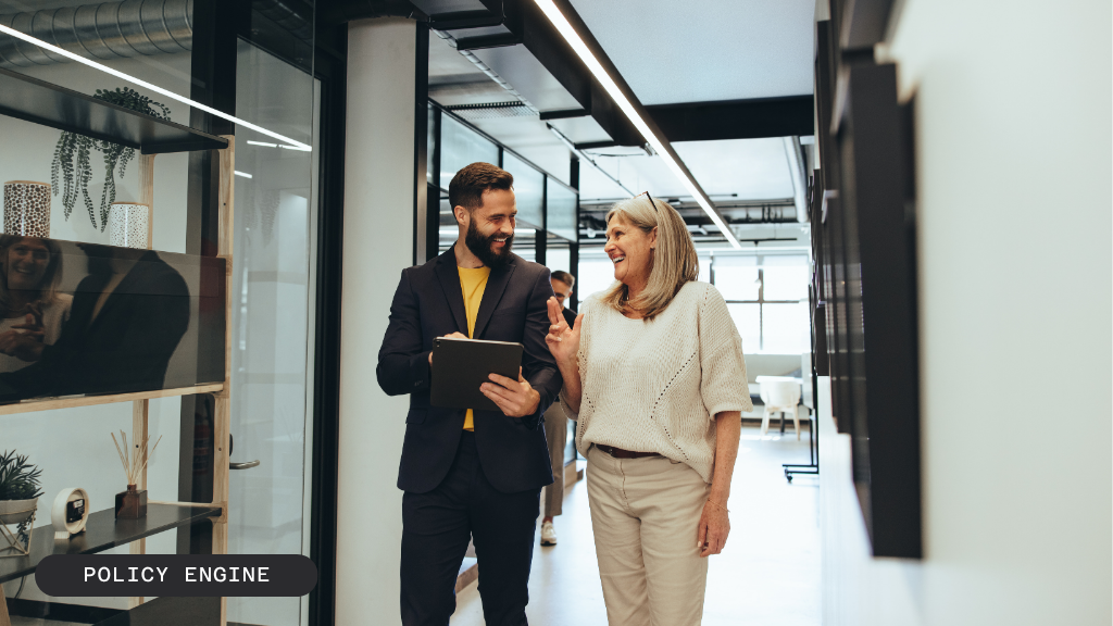 A smiling man holding a tablet walks and talks with an older woman in an office hallway.