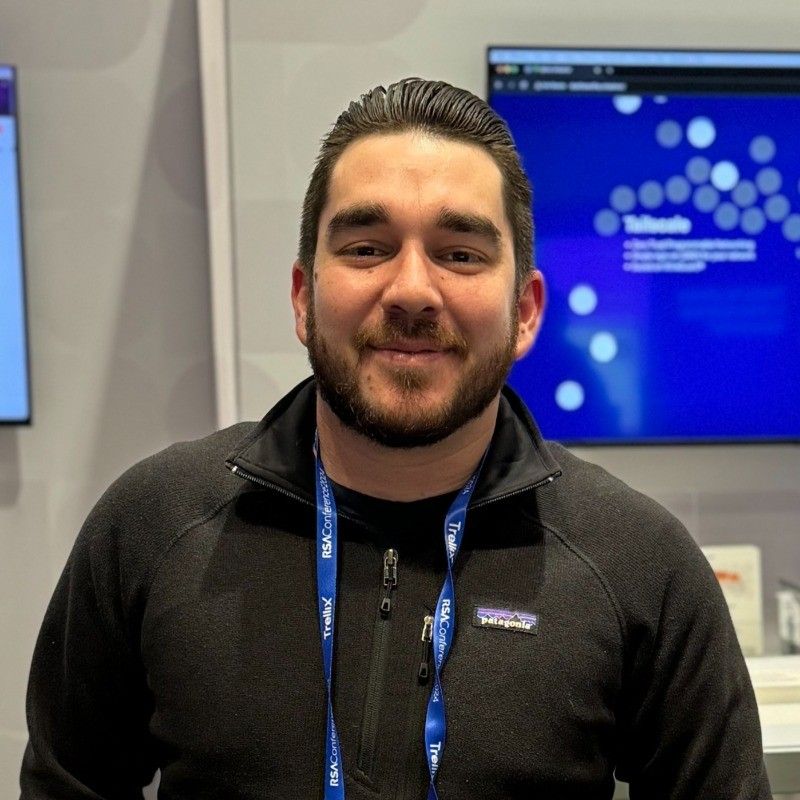 A person with a beard and short hair wearing a black jacket and a blue lanyard stands in front of a display screen with a blue background.
