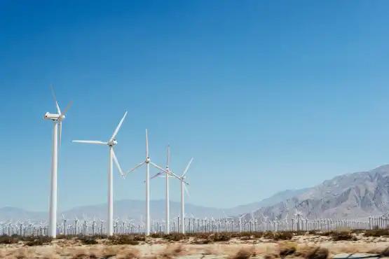 Wind turbines stand in a desert landscape under a clear blue sky, with mountains visible in the background.