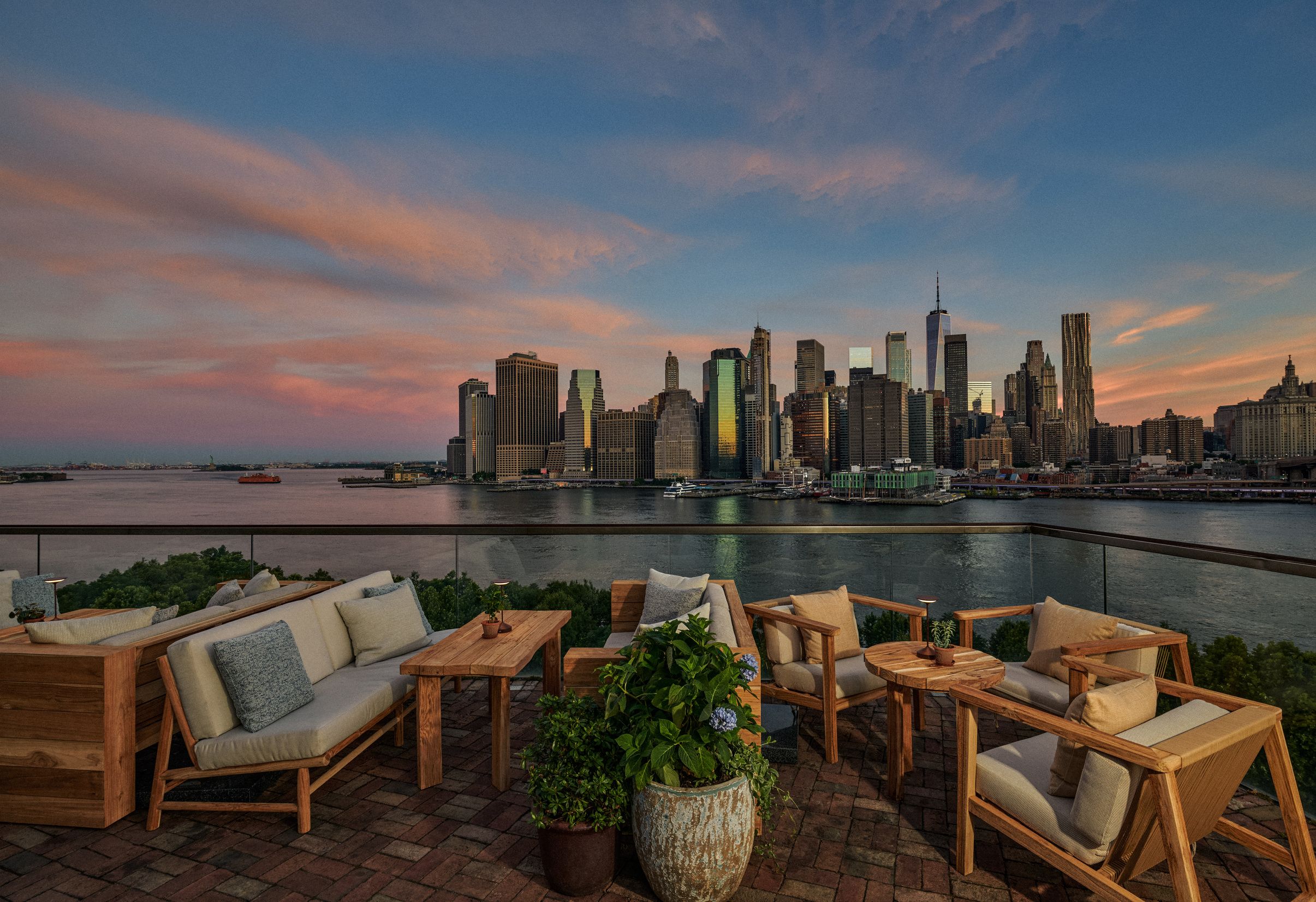Rooftop patio with wooden lounge furniture and potted plants overlooks a city skyline and river during a sunset, featuring a vibrant sky with pink and orange hues.