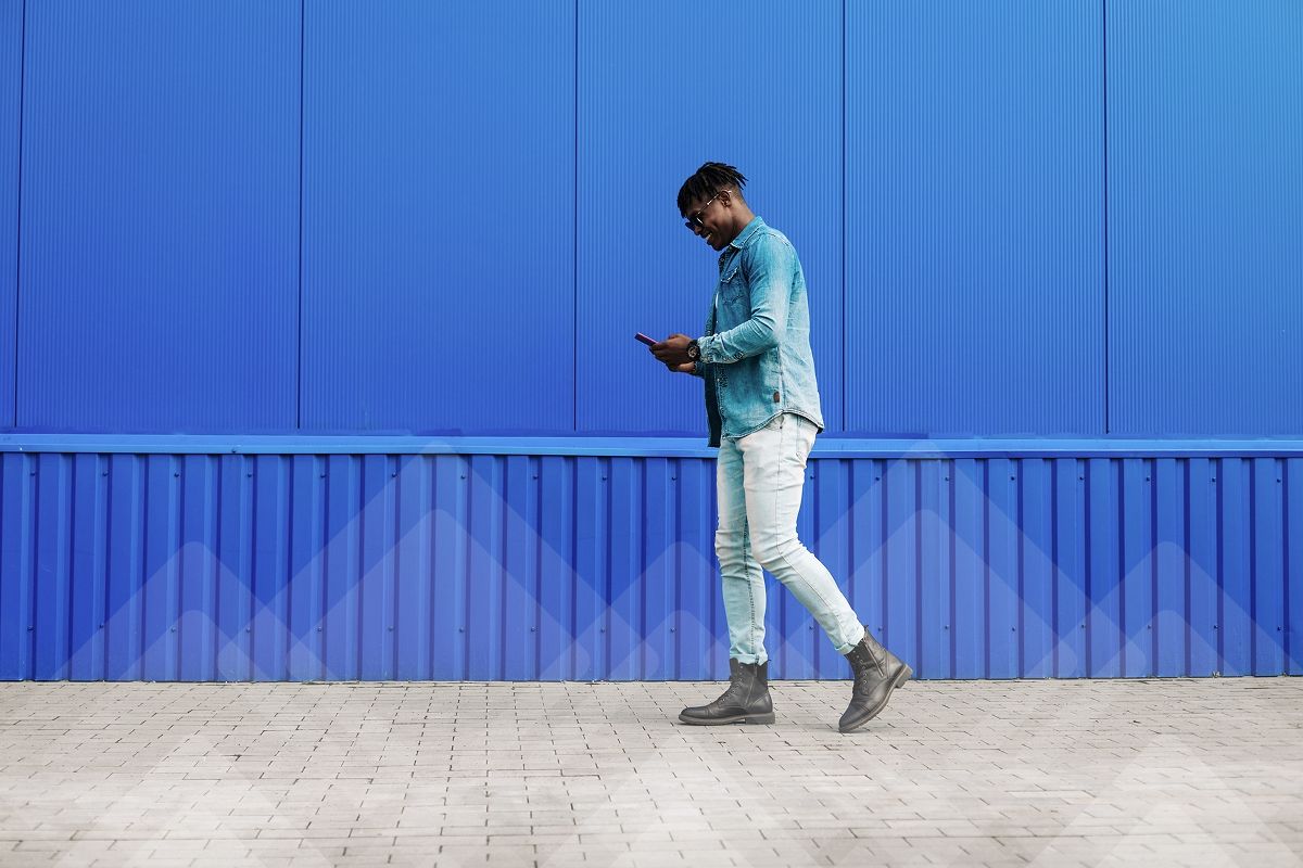 A smiling Black man in a denim jacket and jeans walks past a blue wall while looking at his phone.