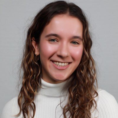 A person with long, wavy brown hair smiles while wearing a white turtleneck against a neutral background.