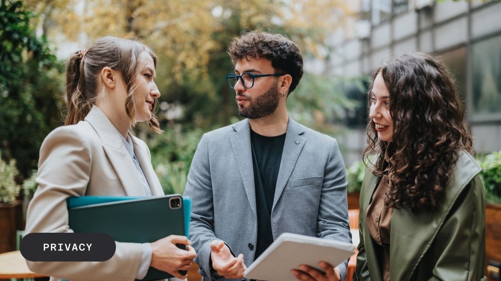 A man with glasses and two women discussing outdoors, holding tablets and a folder.