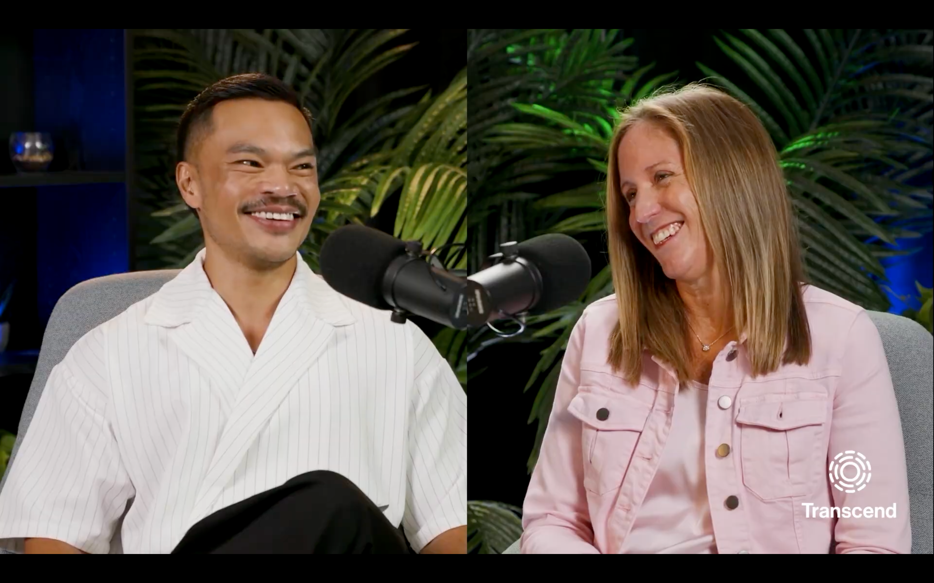 Two people sit in a studio setting with tropical plants, smiling as they speak into microphones. The man on the left wears a white shirt, while the woman on the right wears a pink jacket.