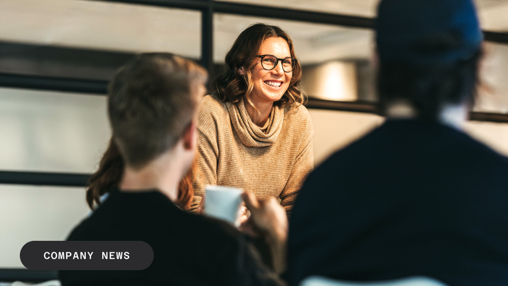 A smiling woman in glasses and a beige sweater looks at two colleagues during a meeting.