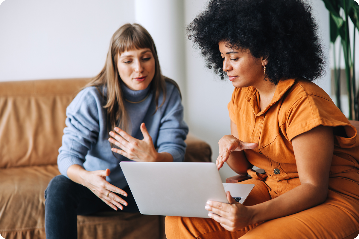 Two women sitting on different couches looking at a single laptop and talking