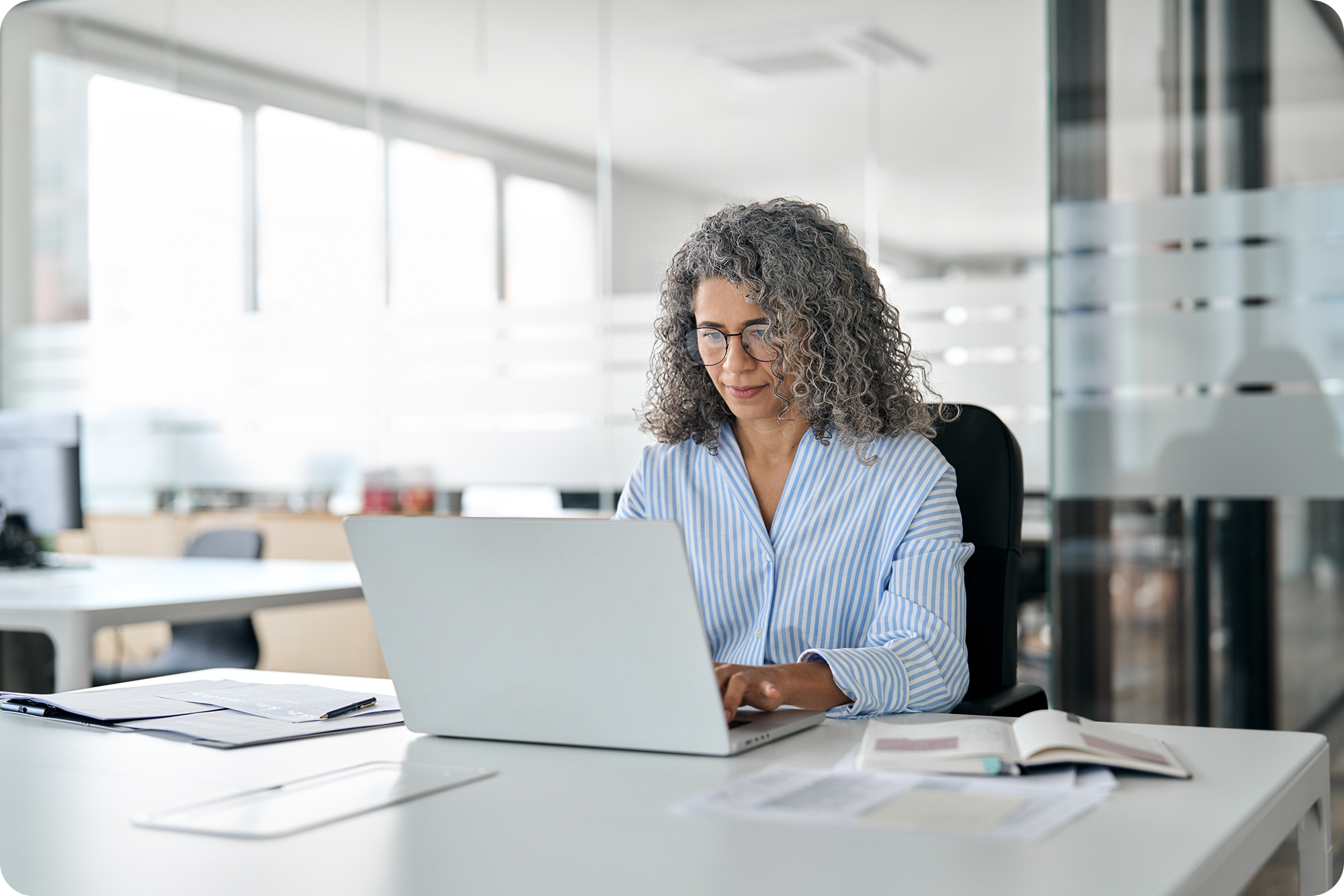 A woman with curly gray hair and glasses is sitting at a desk in an office, working on a laptop. She is wearing a light blue striped blouse, with papers and an open book on the desk.