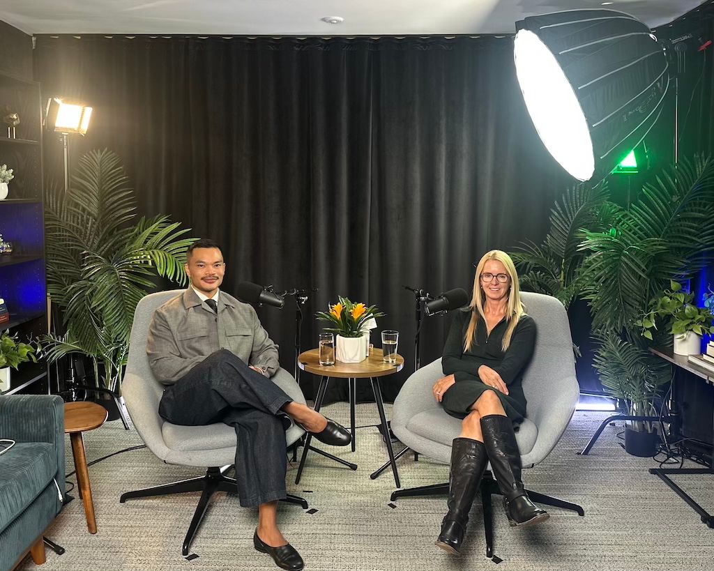 Two people are seated in a studio setup with a dark curtain backdrop, each on a gray chair, surrounded by greenery and studio lighting. They have microphones in front of them and a small table with a flower arrangement and glasses of water between them.