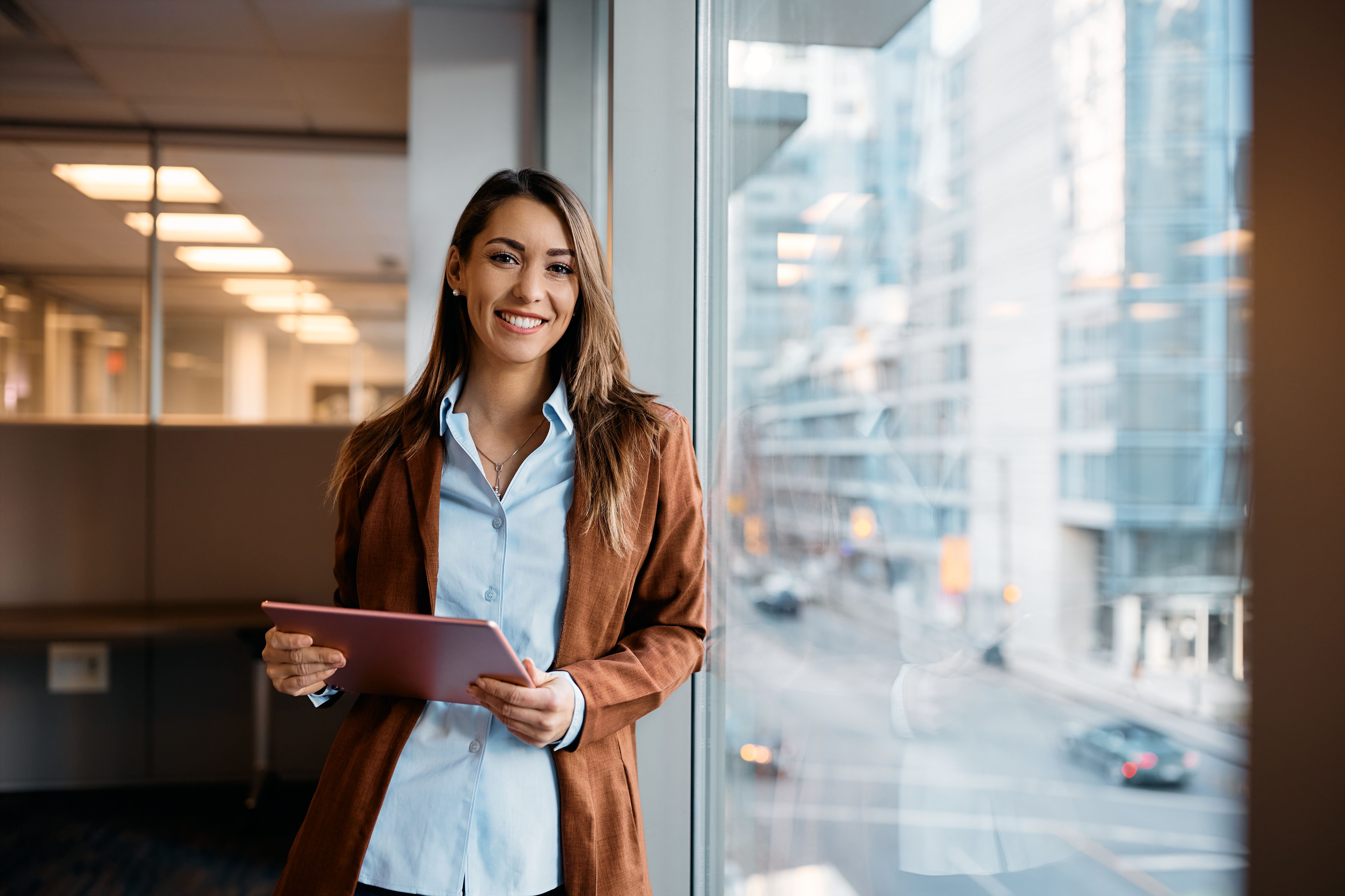 Smiling professional woman holding a tablet in an office with a city view.