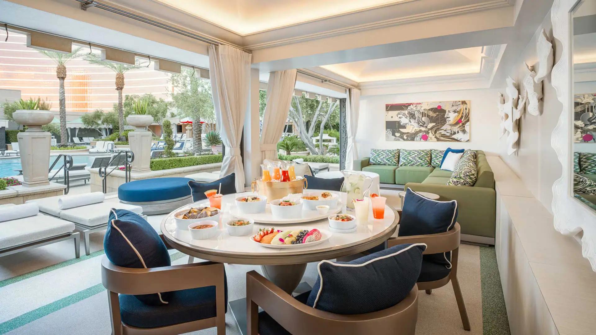 A luxurious poolside cabana features a round table set with a variety of fresh fruits and drinks, surrounded by comfortable seating, blue-cushioned chairs, and an outdoor sofa with decorative pillows. In the background, lush greenery and lounge chairs by the pool are visible through large open windows.