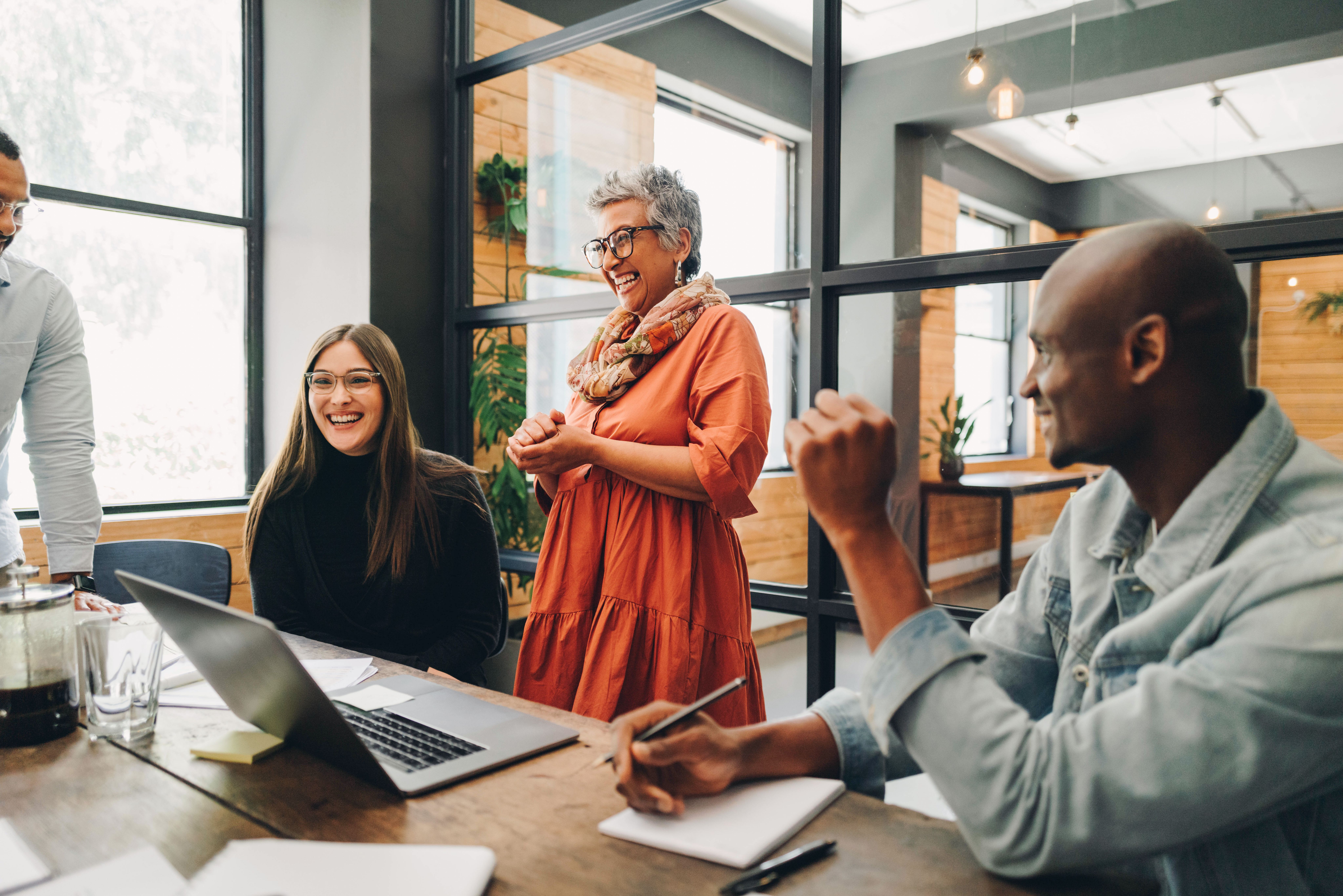 A group of diverse colleagues gather around a table in a modern office, with a woman in an orange dress smiling and speaking, while others listen and take notes. Laptops and notebooks are on the table, creating a collaborative atmosphere.