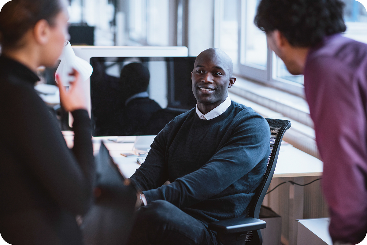 Three colleagues engaged in conversation in a modern office, with one seated at a desk and two standing nearby. A computer monitor is visible in the background.