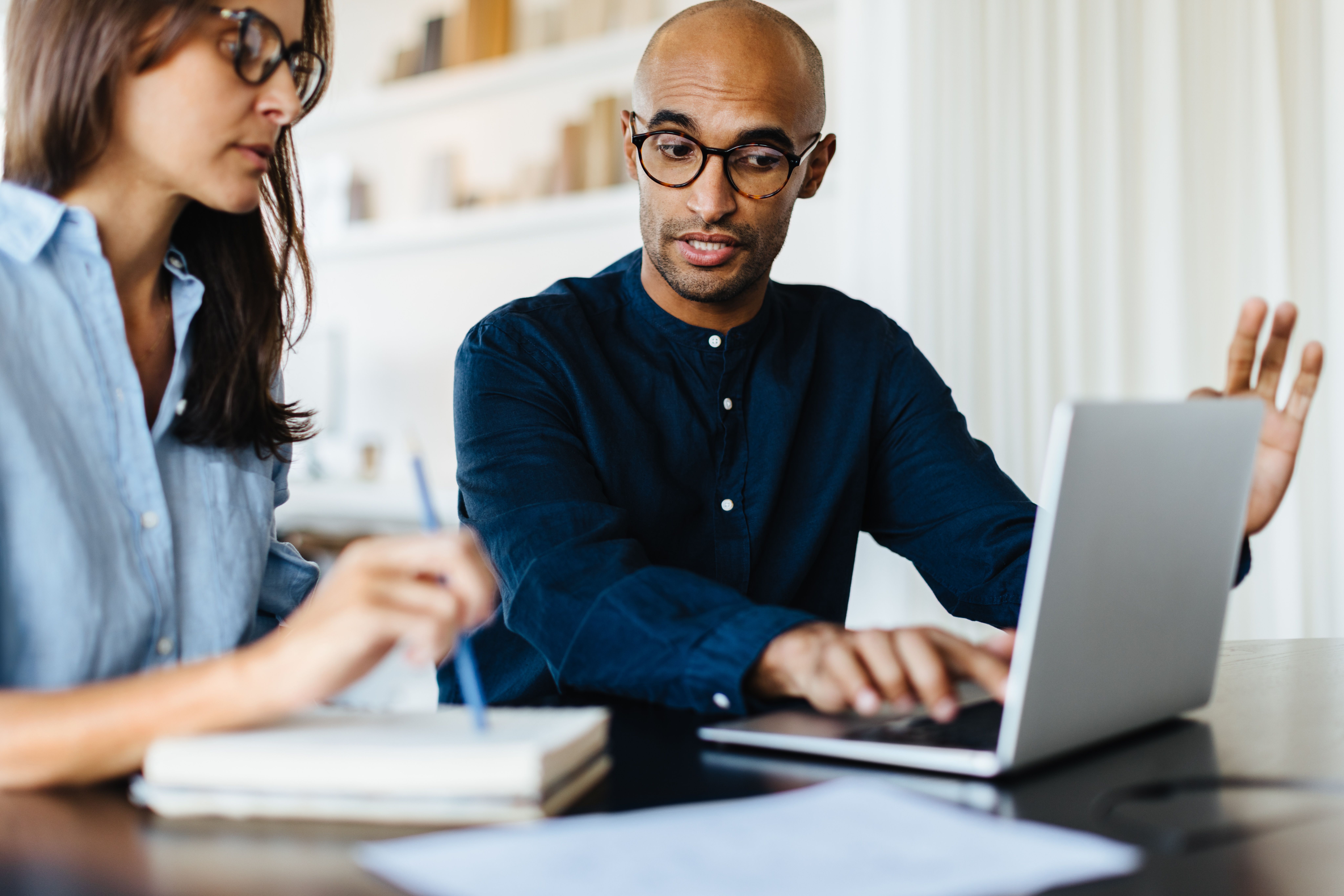 Two people working together at a table, with one person using a laptop and gesturing, while the other takes notes.