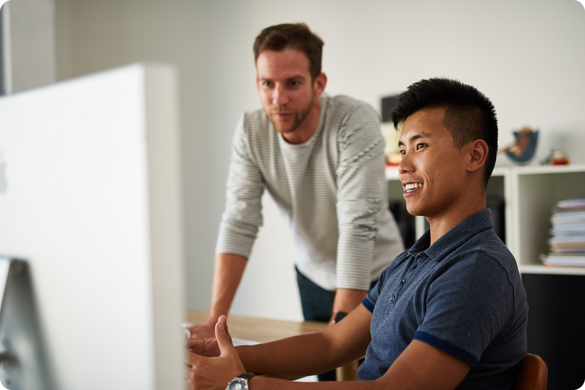 Two men looking at a desktop computer, one is sitting and smiling and the other is standing leaned over the desk