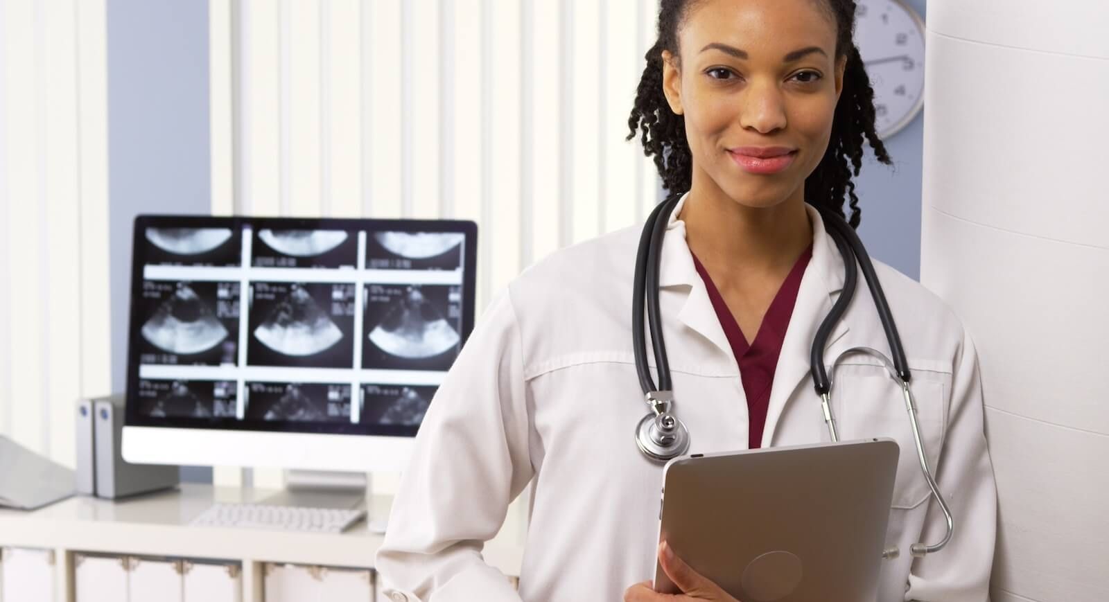 Doctor standing in front of computer screen with medical imaging, holding an ipad