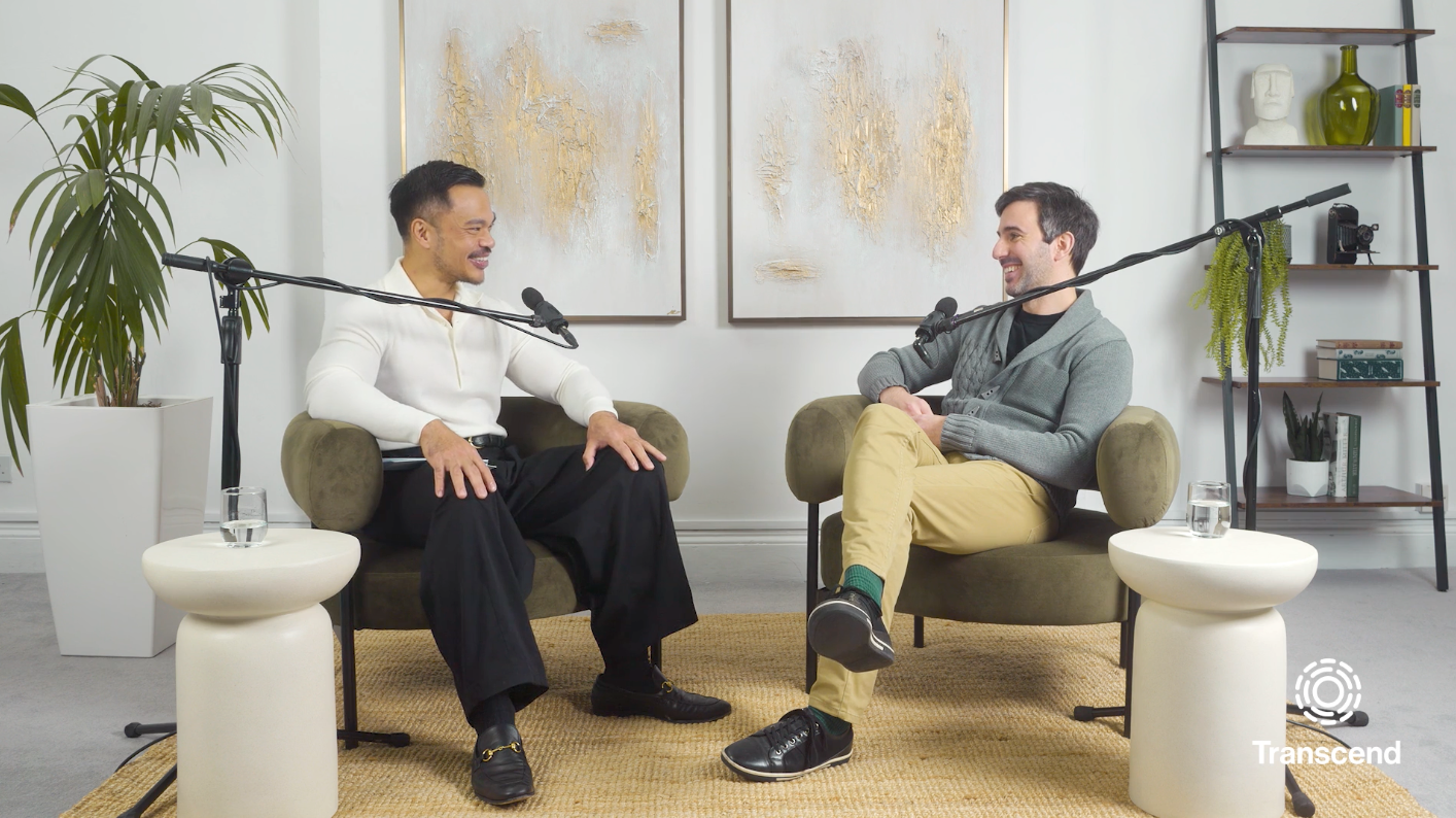Two people sit in armchairs with microphones, engaged in conversation on a beige rug. A potted plant and abstract paintings are in the background, alongside the Transcend logo.