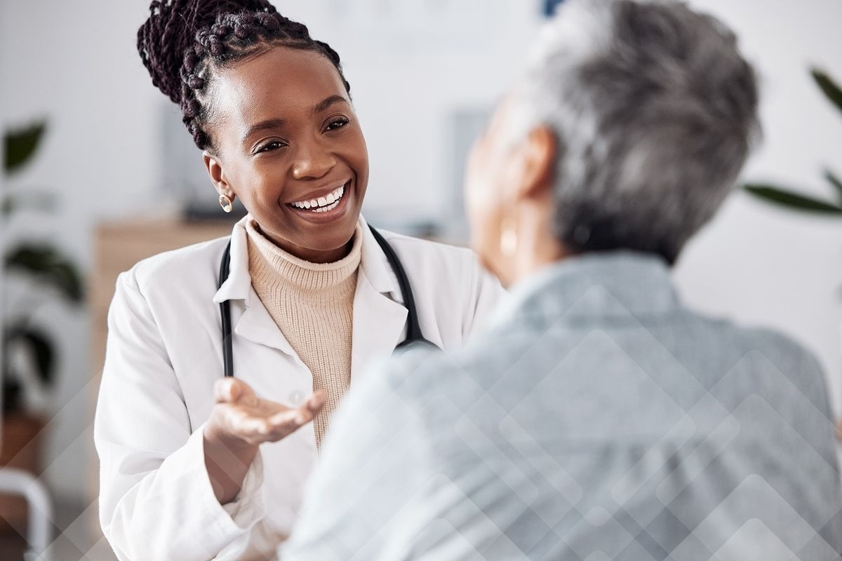 A smiling Black female doctor in a lab coat and stethoscope talks to a patient.