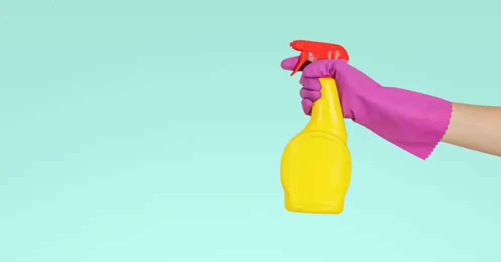 Hand wearing a fushcia cleaning glove and holding a yellow spray bottle over a teal background