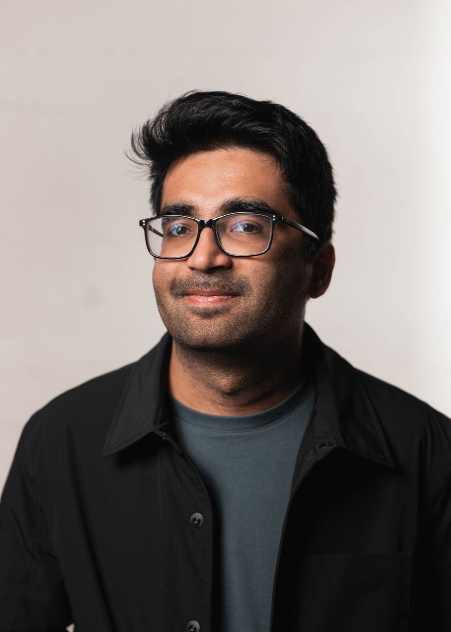 A person with short, dark hair and glasses wearing a dark jacket over a blue shirt, set against a plain background.