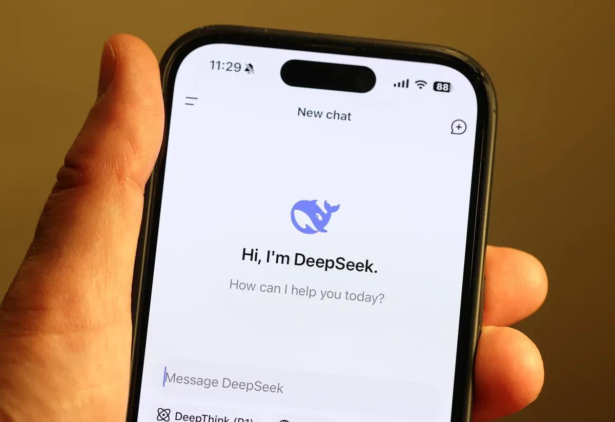 A hand holding a smartphone displaying a chat interface with a blue robot icon and the text "Hi, I'm DeepSeek. How can I help you today?" at the top of the screen.