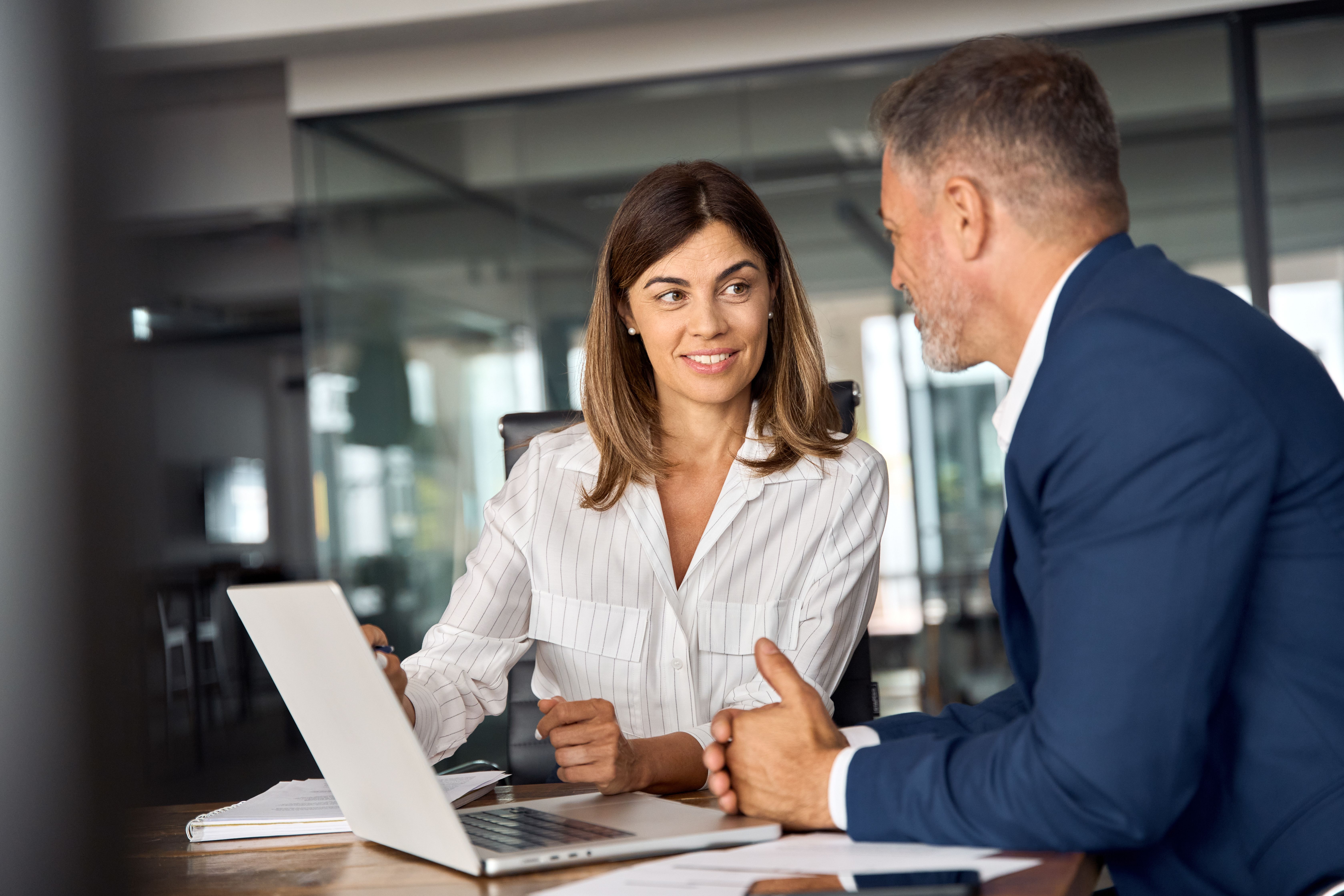 A woman and a man in business attire are sitting at a table in an office, engaged in conversation while looking at a laptop screen.